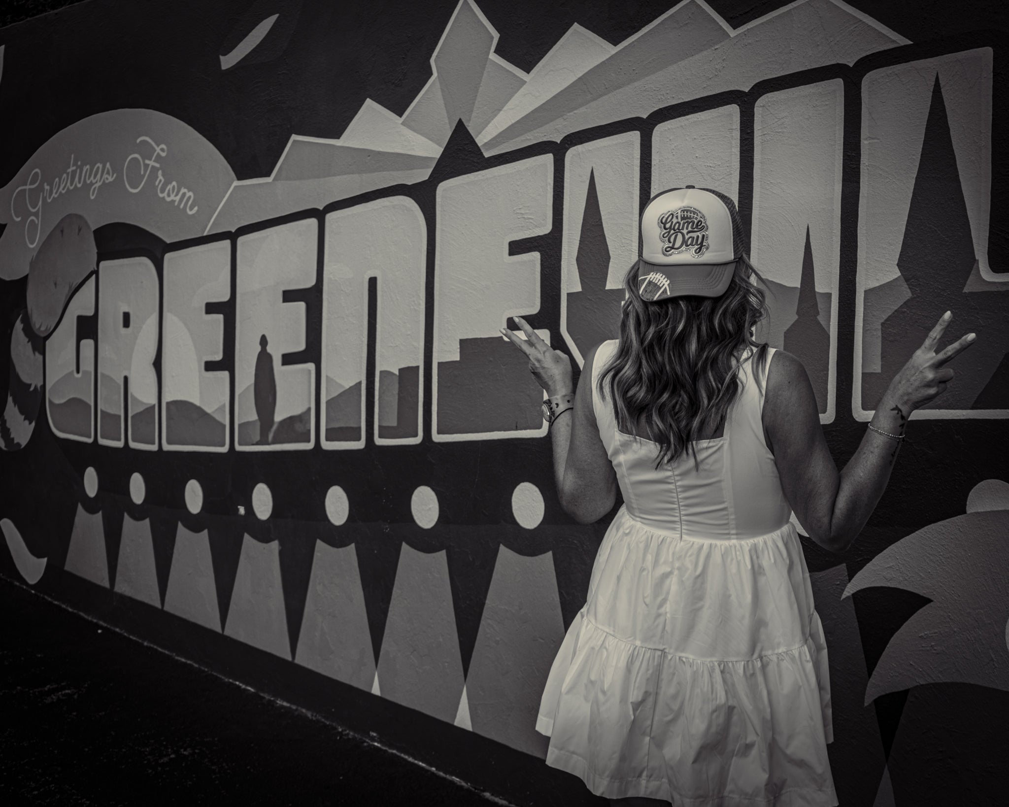 Woman in a white dress and cap standing in front of a 'Greetings from Greenville' mural.